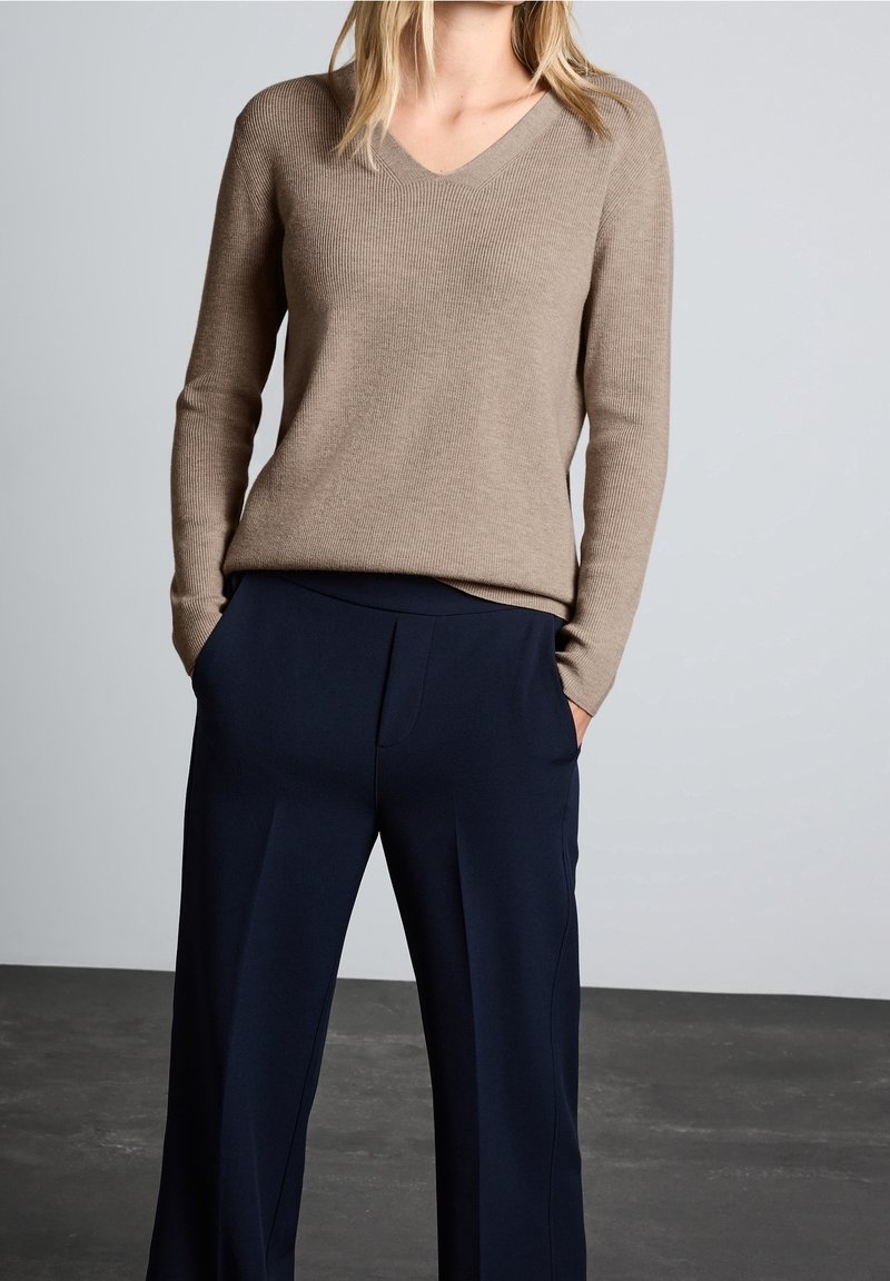 Femme portant un pull beige à col en V et un pantalon large bleu marine, les mains dans les poches, debout devant un fond gris uni.