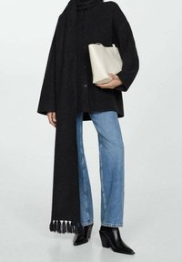 Czarny, oversize'owy płaszcz z wełny z długim szalikiem, zestawiony z jasnoniebieskimi dżinsami w fasonie dzwonów i błyszczącymi czarnymi botkami za kostkę. Trzyma małą, kremową torebkę.