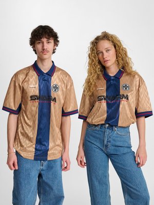 Twee jonge volwassenen dragen bijpassende gouden en marineblauwe sportshirts met "SWARM"-logo's, gecombineerd met een blauwe jeans, staand tegen een effen achtergrond.