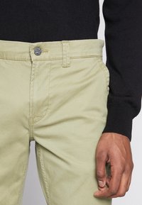 Light green trousers made of cotton blend fabric, featuring a metal button closure, belt loops, and invisible side pockets.