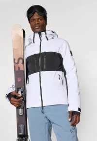 Σακάκι σκι σε λευκό και μαύρο με φερμουάρ, συνδυασμένο με μπλε παντελόνι. Στην μία πλευρά κρατάει ένα σκι με σχέδιο.