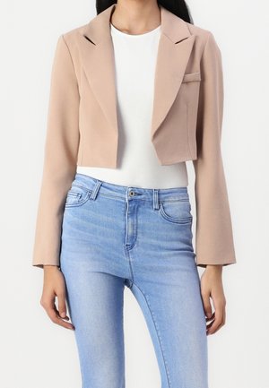 Vrouw die een cropped beige blazer draagt over een wit topje, gecombineerd met lichtblauwe high-waisted jeans, staand tegen een effen achtergrond.