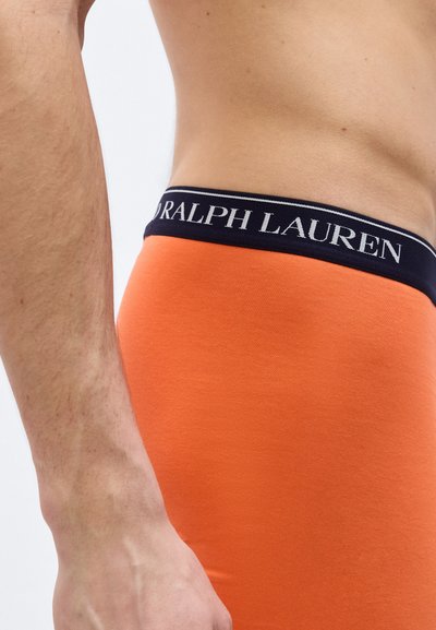 Άνδρας με κορμό και χέρι  που φοράει πορτοκαλί εσώρουχο με μαύρη μέση που φέρει την ετικέτα «Ralph Lauren», πάνω σε απλό λευκό φόντο.