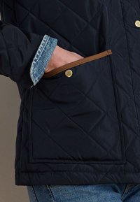 Roka tumši zila dūrainā mētelī ar brūnu apdari un zelta pogu, redzama džinsa krekla aproce, saskaņota ar ziliem džinsiem.