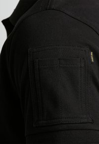 Polo noir en tissu texturé, avec un col en biais et une poche patte sur la manche supérieure avec des détails de couture visibles.