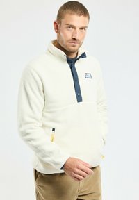 Pull-over en polaire blanche avec un col haut, un devant zippé bleu marine, et une poche. Présente un logo de montagne et un tirette de zip jaune contrastant.