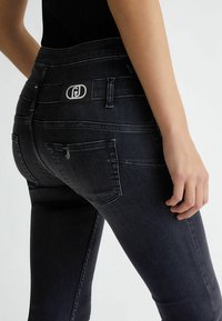 Mujer vistiendo unos jeans ajustados de color azul oscuro con bolsillos traseros y una blusa negra, con la mano relajada a un lado contra un fondo liso.
