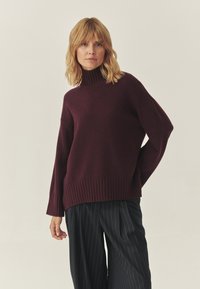 Burgundský sveter s vysokým golierom, ribbed manžetami a lemom. Mäkká textúra, voľný strih, skombinovaný s pruhovanými nohavicami.