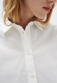 Balta pogu krekla ar klasisku apkakli, izgatavota no gludas auduma ar smalku tekstūru, ar četriem priekšējiem pogām un kārtīgu pogu slīpumu.