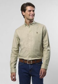 Hellbeiges Hemd mit Knöpfen, langen Ärmeln und Kragen, verziert mit einem kleinen gestickten Logo. Kombiniert mit blauen Jeans und einem braunen Gürtel.