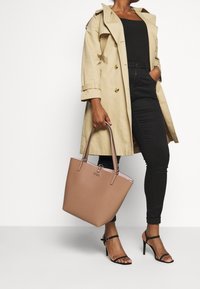 Sac fourre-tout structuré marron clair avec surface texturée et doubles poignées, présentant un logo argenté et un intérieur rose contrastant.