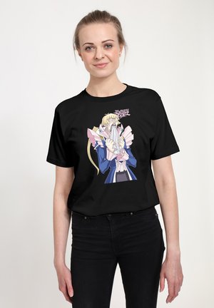 Glimlachende vrouw met blond haar, gekleed in een zwart T-shirt met een animepersonage uit Yu-Gi-Oh! GX, terwijl ze een papier of doek voor haar gezicht houdt.