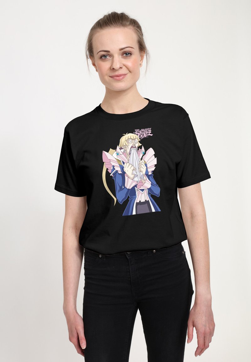 Smiling woman with blonde hair wearing black t-shirt featuring anime character from Yu-Gi-Oh! GX holding a paper or cloth in front of her face.