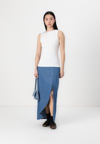 Weißes ärmelloses Oberteil, kombiniert mit einem langen, hellblauen Denimrock mit einem seitlichen Schlitz. Die Accessoires umfassen eine kleine blaue Handtasche und schwarze Schuhe.