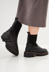 Botas negras hasta el tobillo con paneles laterales elásticos, acabado texturizado, suela gruesa y una tirador. Complementadas con un borde marrón a lo largo de la suela.