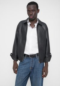 Veste en cuir noire à fermeture éclair avec une coupe décontractée, un large col et des poignets retroussés. Associée à une chemise claire boutonnée et un jean en denim bleu.