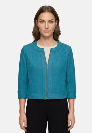 Femme aux cheveux bruns mi-longs portant une veste courte turquoise à manches trois-quarts avec une fermeture éclair dorée, par-dessus un haut blanc et un pantalon noir.