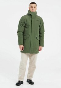 Veste verte isolée avec un col haut et une capuche. Présente deux poches avant et une fermeture éclair. Portée sur un pantalon beige clair et des bottes noires.