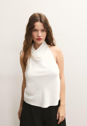 Frau mit langen, welligen Haaren trägt ein ärmelloses weißes Neckholder-Top und schwarze Hose, steht vor einem schlichten, hellen Hintergrund.