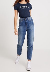 Mörkblå T-shirt med "TOMMY"-logotyp, högmidjade ljusblå jeans och vita sneakers. Rak bendesign. Slät denimtextur.