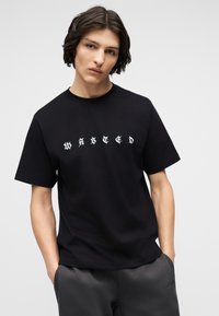 Νεαρός με μεσαίου μήκους σκούρα μαλλιά που φοράει μαύρο μπλουζάκι με λευκή γοτθική επιγραφή "WASTED" και γκρι παντελόνι, με τα χέρια στις τσέπες.