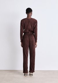 Combinaison à manches longues en rouge foncé avec des rayures métalliques verticales. Taille élastique, coupe ample, et jambes de pantalon évasées. Tissu lisse et texturé.