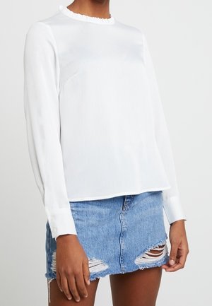 Mujer con blusa blanca de manga larga con cuello con volantes y minifalda vaquera azul claro desgastada.
