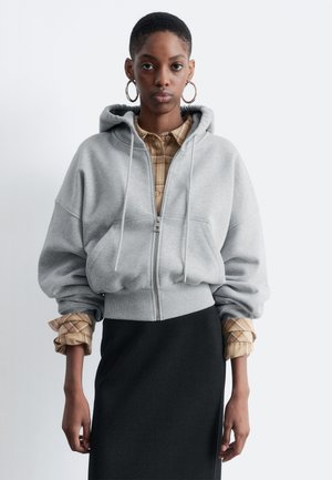 Gray zip-up hoodie with a hood, oversized fit, ribbed hem, and cuffed sleeves. Worn over a beige plaid shirt and black skirt.