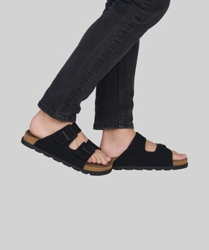 Pieds portant des sandales noires à boucle avec semelles en liège, associées à un jean noir, sur un fond gris uni.