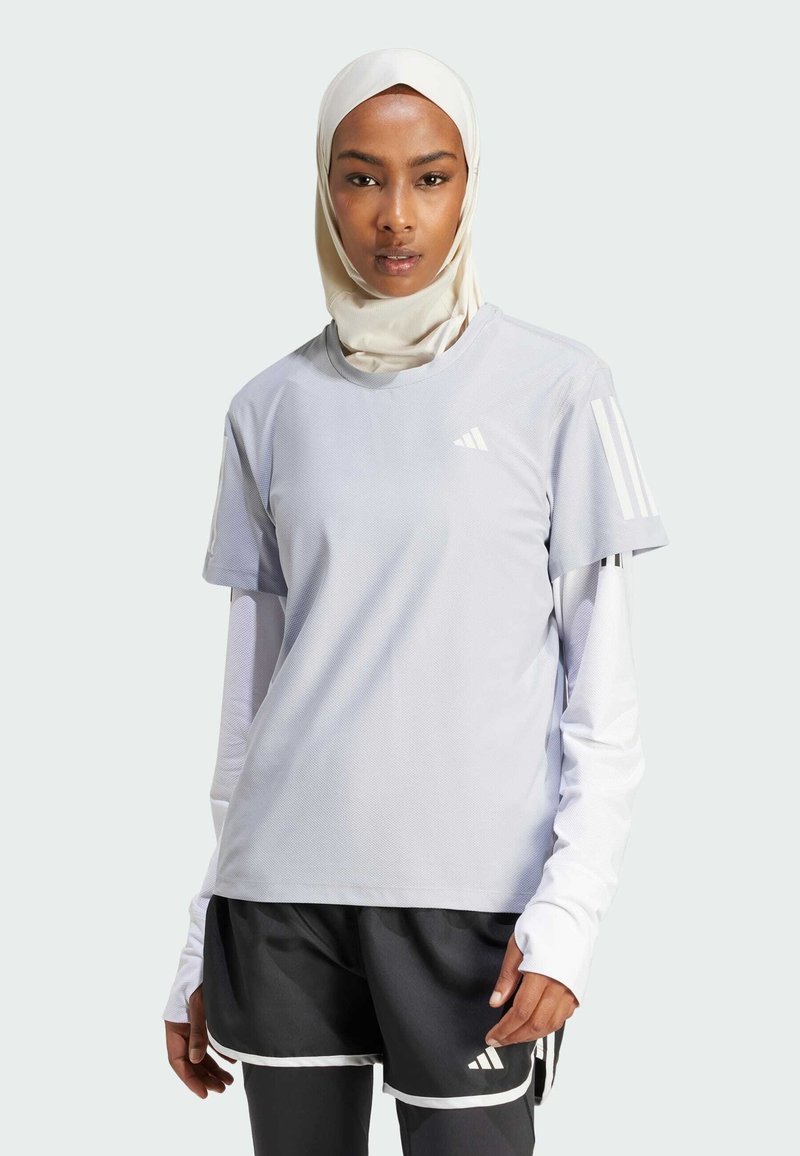 Top deportivo gris claro de manga corta superpuesto sobre una camiseta blanca de manga larga, combinado con pantalones cortos negros con detalles en blanco.