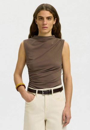 Donna con lunghi capelli castani che indossa un top marrone senza maniche, pantaloni crema, cintura marrone e braccialetti robusti, in piedi con una mano in tasca.