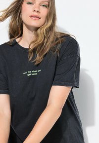 Sort bomulds t-shirt med en afslappet pasform, der har trykt tekst "sms mig når du er hjemme" i lys grøn. Korte ærmer og rund halsudskæring.
