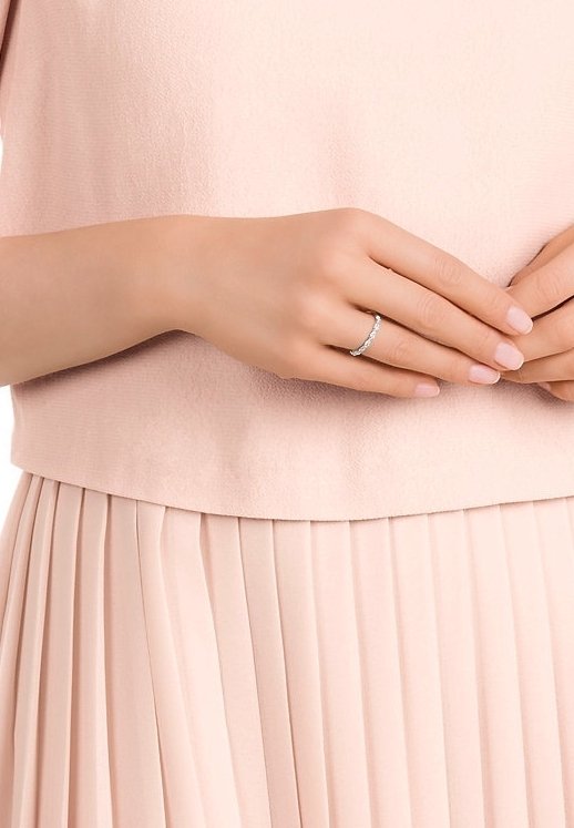 Mains de femme avec une bague en argent à la main gauche, portant un haut rose clair et une jupe plissée, les doigts se touchant doucement.