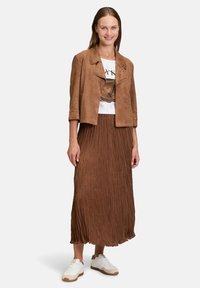 Veste en suède brun, courte, avec des accents cloutés, superposée sur un T-shirt blanc à motifs, associée à une jupe maxi plissée marron et des baskets blanches.
