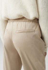 Beige velvet pants with an elastic waistband, featuring back pockets and a smooth texture. Worn with a cream-colored knitted sweater.