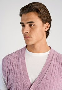 Hellrosa Strickweste mit Zopfmuster, tiefem V-Ausschnitt und gerippten Akzenten, getragen über einem weißen T-Shirt. Glatte Textur und lockere Passform.