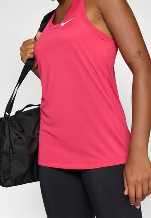 Persoon draagt een roze mouwloos Nike-topje en zwarte leggings, houdt een zwarte sporttas over één schouder met een aardbeitentattoo op de arm.