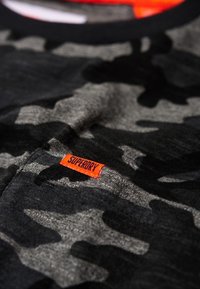 Tričko s kamuflážním potiskem v odstínech černé a šedé, s výraznou oranžovou nálepkou "SUPERDRY" u výstřihu. Měkká textilie.