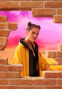 Mujer joven con un peinado de moños espaciales que lleva una chaqueta amarilla, de pie detrás de una pared de ladrillos rota, con un cielo vibrante en tonos rosa y naranja.