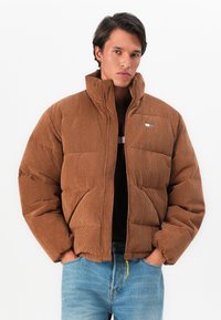 Jonge man in een bruin corduroy pufferjack met hoge kraag, handen in de zakken, gecombineerd met lichtblauwe spijkerbroek tegen een witte achtergrond.