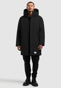 Čierny nepremokavý parka s kapucňou, má predný zips a cvoky, dve bočné vrecká a logo nášivku na leme. Nosené cez čierne nohavice.