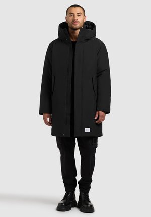 Schwarze wasserdichte Parka mit Kapuze, ausgestattet mit einem Frontreißverschluss und Druckknöpfen, zwei Seitentaschen und einem Logopf patch am Saum. Über schwarzen Hosen getragen.