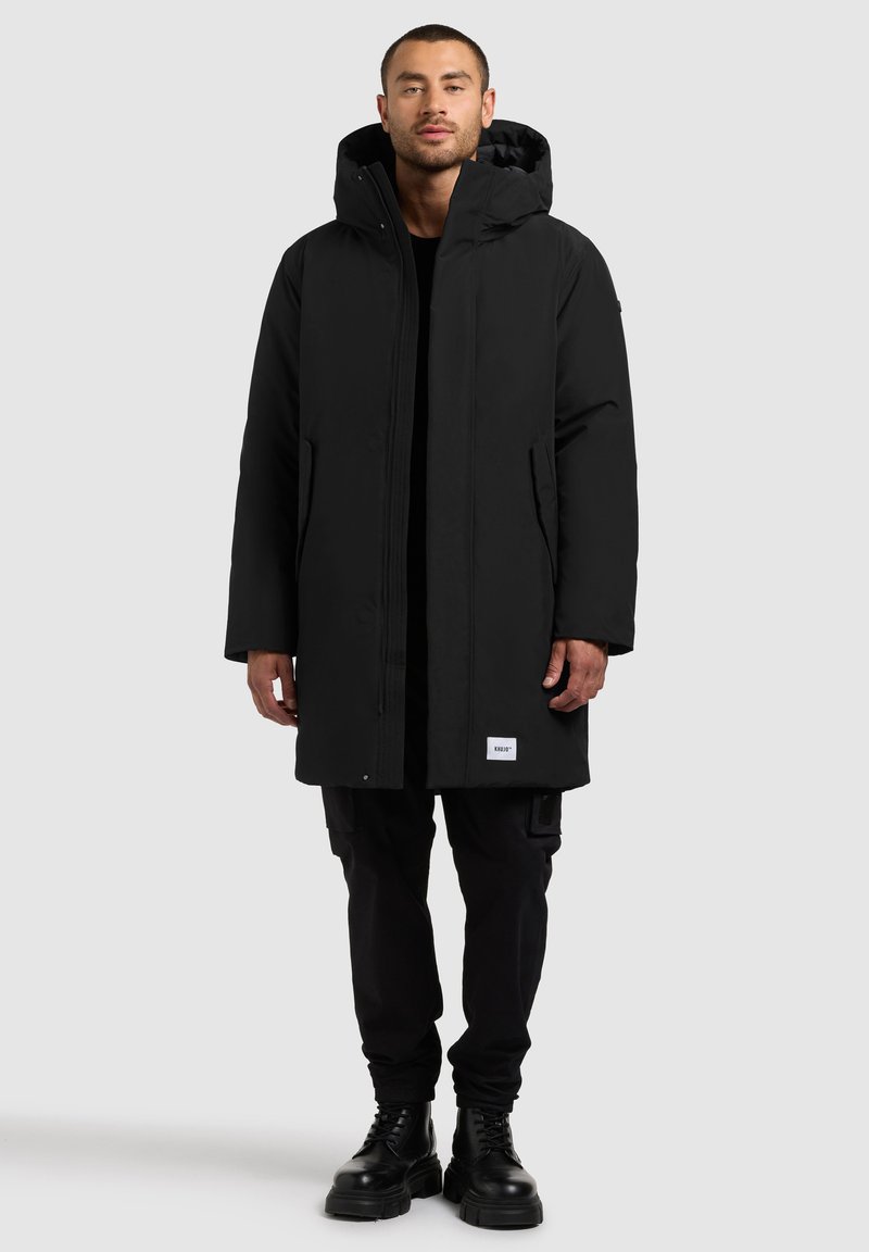 Čierny nepremokavý parka s kapucňou, má predný zips a cvoky, dve bočné vrecká a logo nášivku na leme. Nosené cez čierne nohavice.