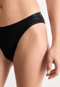 Close-up van een persoon die zwarte merkondergoed in bikini-stijl draagt, waarbij hun heup, onderbuik en hand zichtbaar zijn tegen een witte achtergrond.