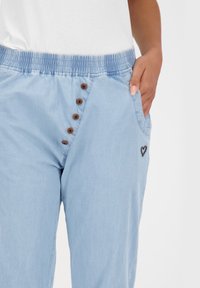 Jasnoniebieskie spodnie z miękkiego materiału, z elastycznym pasem, brązowymi guzikami oraz małą czarną łatą w kształcie serca na kieszeni.