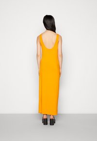 Długa, dopasowana sukienka w jaskrawym pomarańczowym kolorze z dekoltem w serek, wykonana z miękkiego materiału, sięgająca kostek, zestawiona z czarnymi butami na platformie.
