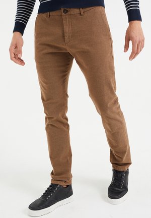 Hombre vistiendo pantalones de pana marrones, zapatillas negras con suelas blancas y un suéter azul marino con rayas blancas en las mangas, de pie sobre un suelo blanco.