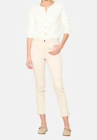 Vit texturerad cardigan med mässingsknappar, kombinerad med ljusbeige åtsittande byxor och ljusbruna skor med dekorativa utskärningar.