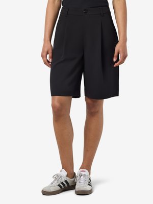 Person trägt schwarze kniehohe, maßgeschneiderte Shorts und weiße Adidas-Sneaker mit schwarzen Streifen, steht vor einem weißen Hintergrund.