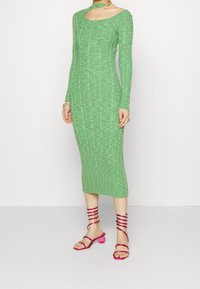Abito a maglia verde a coste con maniche lunghe e scollatura ampia. Abbinato a sandali con tacco rosa con cinturini a spirale e accenti trasparenti.